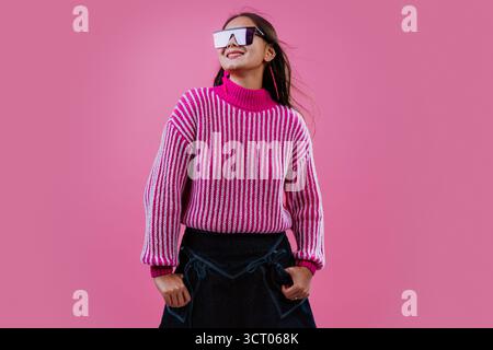 Eine junge Frau lächelt, trägt einen rosa gestreiften Pullover, eine herzförmige Sonnenbrille und blitzschnelle Ohrringe und macht eine Rock-and-Roll-Handgeste gegen Stockfoto