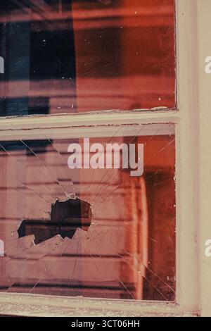 Eine kaputte Fensterscheibe mit Spinnennetz-ähnlichen Rissen und warmen Reflexen. Die Szene weckt ein Gefühl von Verlassenheit, Unvollkommenheit und stiller Spannung. Stockfoto
