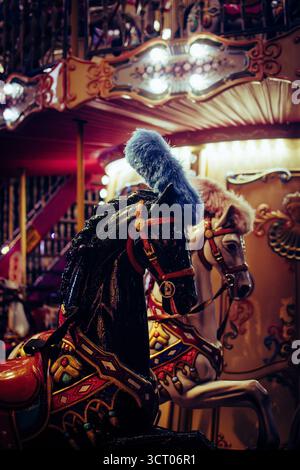 Nächtliche Nahaufnahme von verzierten Karussellpferden in Łódź, Polen, deren detailreiche Sättel und gefiederte Kopfstücke unter warmen Karnevalslichtern leuchten. Stockfoto