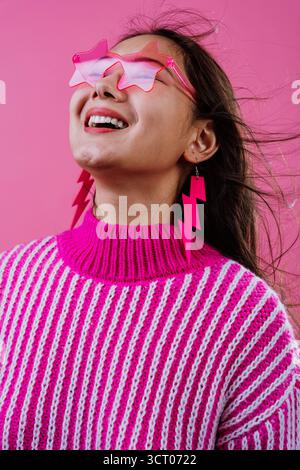 Junge Frau lächelt mit geschlossenen Augen in leuchtend rosa sternenförmigen Brillen, Blitzohrringen und gestreiftem Strickpullover vor farblich passenden Ba Stockfoto