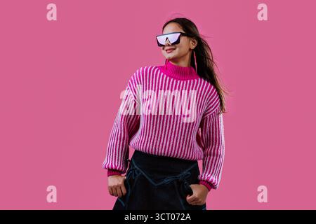 Eine junge Frau lächelt, trägt einen rosa gestreiften Pullover, eine herzförmige Sonnenbrille und blitzschnelle Ohrringe und macht eine Rock-and-Roll-Handgeste gegen Stockfoto