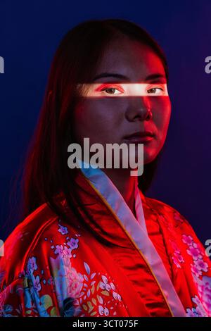 Asiatische Frau posiert in einem traditionellen roten Kimono mit floralen Mustern, steht vor einem dunkelblauen Hintergrund mit dramatischem rotem Licht, das einen erhellt Stockfoto