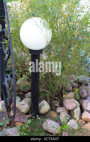 Ein Garten mit Beleuchtung, ausgestattet mit elektrischen Globus-Lampen, in einem offenen Landschaftsgarten. Die Lampe an einem Pfosten ist vor dem Hintergrund eines grünen Bu montiert Stockfoto