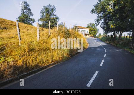 Kurvige Landstraße führt bergauf durch goldenes Herbstgras und Bäume, mit einem kleinen Haus auf der Spitze unter klarem blauem Himmel, friedliche Landschaft Stockfoto