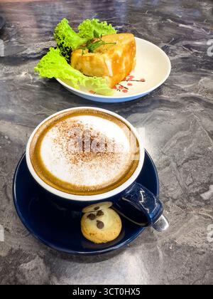 Ein schaumiger Cappuccino mit Zimt, serviert in einer blauen Tasse neben einem kleinen Keks auf einem Marmortisch mit einem kaya Toast im Hintergrund malaysia Stockfoto