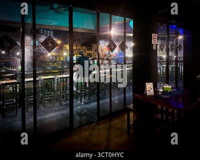 Eine Einzelfigur sitzt an einer Bar in einem schwach beleuchteten Gebäude mit Blick auf eine lebhafte Stadtstraße durch große Glasfenster Stockfoto