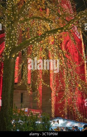 Ein wunderschön beleuchteter Baum mit warmen Weihnachtslichtern steht vor dem rot beleuchteten Tempel Saint-Etienne auf dem Weihnachtsmarkt in Mulhouse in Frankreich. Stockfoto