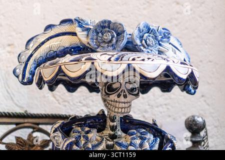 Ein blau-weißer Keramikschädel mit Blumen darauf. Der Schädel wird auf einen Tisch gestellt und die Blumen sind um ihn herum angeordnet. Die Szene ist unheimlich und so Stockfoto