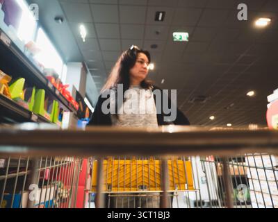 Lombardei, Italien - 1. Oktober 2025 Eine Frau mit langen dunklen Haaren, die einen grauen Pullover und eine schwarze Strickjacke trägt, schiebt einen Einkaufswagen durch eine hell beleuchtete Stockfoto