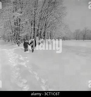 Ein atmosphärisches Schwarz-weiß-Archivfoto aus den 1970er Jahren, das einen ruhigen Winterspaziergang in der Mayaky Forestry, der Ukrainischen SSR, aufnimmt. Mutter und Kind schlendern gemeinsam durch eine riesige Landschaft aus tiefem, frischem Schnee und hinterlassen Spuren. Sie laufen am Rande eines schneebedeckten Laubwaldes entlang, dessen Äste zart frostig sind. Dieses ruhige und minimalistische Bild ist eine nostalgische Darstellung eines friedlichen Familienausflugs und der einfachen Freuden einer sowjetischen Kindheit im Winter Stockfoto