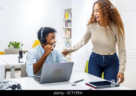Multiethnische Kollegen lächeln und interagieren in einem modernen Büro, während ein Mann ein Headset trägt und sein Laptop benutzt, während eine Frau in der Nähe steht und sich darüber diskutiert Stockfoto