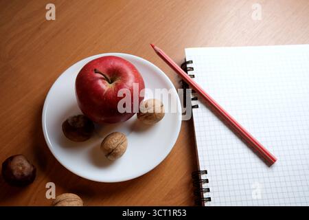 Ein hellroter Apfel mit Walnüssen sitzt auf einem Teller neben einem Notizblock und Bleistift. Stockfoto