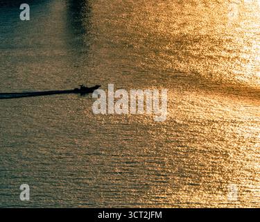 Ein einsames Boot gleitet über das goldene Meer von Mokpo, Südkorea, und hinterlässt ein sanftes Licht, während sich das Licht des Sonnenuntergangs auf dem ruhigen, glitzernden Wasser reflektiert. Stockfoto