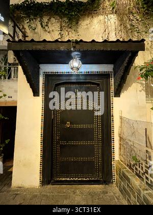 Verzierte schwarze Tür mit goldenen Nieten, Fliesenrahmen und Laterne-Glühen – markiert mit „15 Rue Tree“. Tanger, Marokko. 4. Oktober 2025. Stockfoto