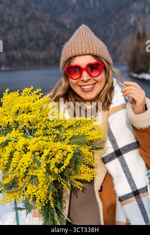 Mimosa Flowers Woman Winter Mountains: Glückliche lächelnde Frau mit Mimosa-Blumenstrauß in der Nähe des Sees, die den Frühling in der Berglandschaft feiert. Stockfoto