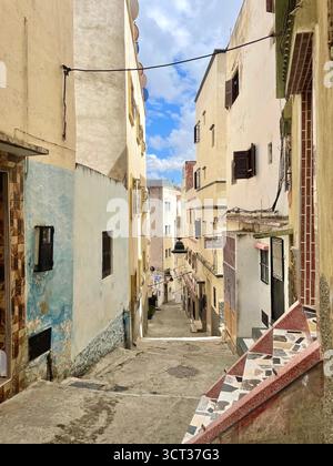 Schräge Gasse in der Alten Medina mit mehrschichtigen Fassaden, gekachelten Stufen und einer Mischung aus alten Texturen unter teilweise bewölktem Himmel. Tanger, 7. Juni 2024 Stockfoto