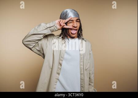 Ein junger Mann mit langen Haaren, der ein Bandana und ein leichtes Hemd trägt, schlägt eine lustige Pose, streckt seine Zunge aus und macht in einem einfachen Studi ein Friedenszeichen Stockfoto