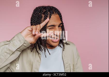 Eine junge Person zeigt einen fröhlichen Ausdruck, während sie mit einer Hand ein Friedenszeichen macht. Die verspielte Pose wird vor einem sanften rosa Hintergrund inszeniert Stockfoto