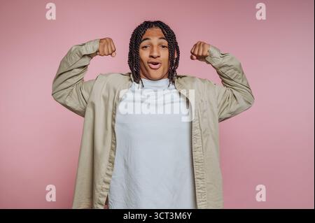 Ein junger Mensch mit geflochtenen Haaren lächelt weit, während er seinen Bizeps beugt. Sie tragen ein weiches weißes Hemd und stehen vor einem zarten rosa B Stockfoto