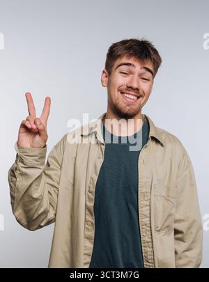 Ein junger Mann steht vor einem hellen Hintergrund, lächelt weit und zeigt mit den Fingern ein Friedenszeichen. Er trägt ein lässiges Hemd über einem Schweiß Stockfoto