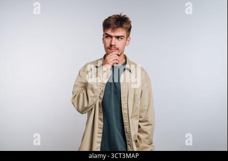 Ein junger Mann steht in einem hell beleuchteten Studio und trägt ein lässiges Outfit. Er hat einen nachdenklichen Ausdruck, als er sein Kinn berührt, verloren in der Nachdenklichkeit abo Stockfoto