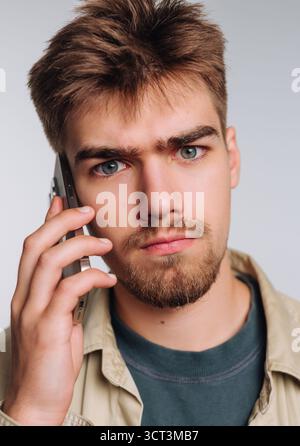 Ein junger Mann mit hellem Bart und durchdringenden blauen Augen spricht intensiv auf einem Smartphone. Er trägt ein lässiges Hemd und wirkt fokussiert, gegen eine Keule gestellt Stockfoto