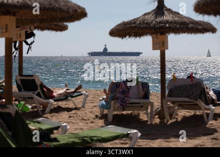 Palma, Spanien. Oktober 2025. Die Menschen sonnen sich am Strand von Can Pere Antoni, während der US-Flugzeugträger USS Gerald R. Ford in der Bucht von Palma vor Anker liegt. Das größte und modernste Kriegsschiff der US Navy ist gestern auf Mallorca angekommen und soll bis zum 8. Oktober bleiben. Quelle: Clara Margais/dpa/Alamy Live News Stockfoto