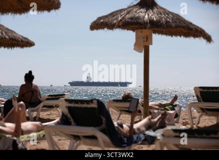 Palma, Spanien. Oktober 2025. Die Menschen sitzen am Strand von Can Pere Antoni, während der US-Flugzeugträger USS Gerald R. Ford in der Bucht von Palma vor Anker liegt. Das größte und modernste Kriegsschiff der US Navy ist gestern auf Mallorca angekommen und soll bis zum 8. Oktober bleiben. Quelle: Clara Margais/dpa/Alamy Live News Stockfoto