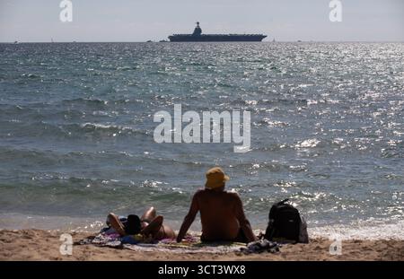 Palma, Spanien. Oktober 2025. Die Menschen sonnen sich am Strand von Can Pere Antoni, während der US-Flugzeugträger USS Gerald R. Ford in der Bucht von Palma vor Anker liegt. Das größte und modernste Kriegsschiff der US Navy ist gestern auf Mallorca angekommen und soll bis zum 8. Oktober bleiben. Quelle: Clara Margais/dpa/Alamy Live News Stockfoto