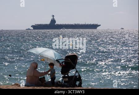 Palma, Spanien. Oktober 2025. Die Menschen sitzen am Strand von Can Pere Antoni, während der US-Flugzeugträger USS Gerald R. Ford in der Bucht von Palma vor Anker liegt. Das größte und modernste Kriegsschiff der US Navy ist gestern auf Mallorca angekommen und soll bis zum 8. Oktober bleiben. Quelle: Clara Margais/dpa/Alamy Live News Stockfoto