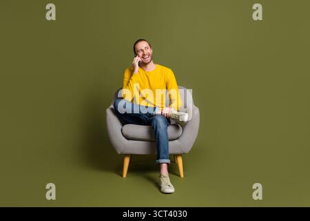 Junger Mann, der auf einem Stuhl sitzt und auf einem Smartphone spricht, während er sich fröhlich anspricht, vor einem khakifarbenen Hintergrund Stockfoto