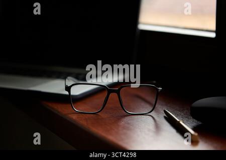 Brille mit Stift auf einer polierten Schreibtischoberfläche neben dem Laptop mit verschwommenem Hintergrund. Saubere professionelle Szene für Business-Lifestyle-Konzepte. Stockfoto