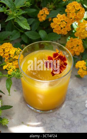 Ein erfrischender gelb-oranger tropischer Cocktail, serviert in einem klaren Glas mit einem großen Eiswürfel, garniert mit einer leuchtenden roten und orangen Ringelblume. Stockfoto