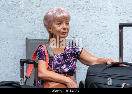 Eine ältere Frau mit einem kurzen Haarschnitt und einer Tasche sitzt neben großen Koffern Stockfoto