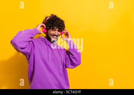 Fröhlicher Mann in einem stilvollen lila Hoodie, der Kopfhörer trägt, lächelt und Musik auf einem leuchtend gelben Hintergrund genießt Stockfoto