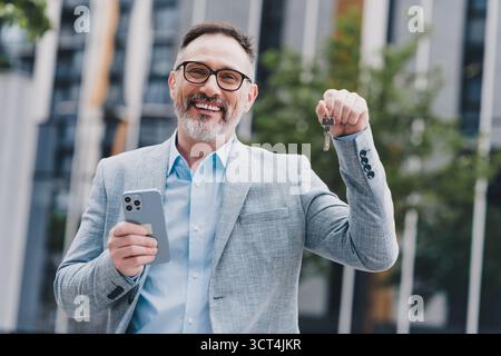 Ein Geschäftsmann in einer modernen Stadt lächelt, während er Schlüssel und ein Smartphone in einer urbanen Straßenszene hält, die Selbstvertrauen und Erfolg zeigt Stockfoto
