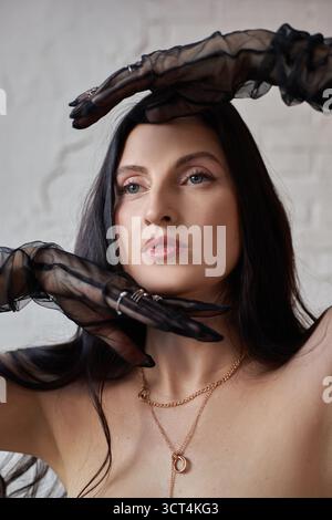 Das Model zeigt eine auffällige Pose mit anmutig positionierten Armen und trägt durchsichtige schwarze Handschuhe, die einen Kontrast zu ihrem langen Haar bilden. Die Einstellung ist einfach wi Stockfoto