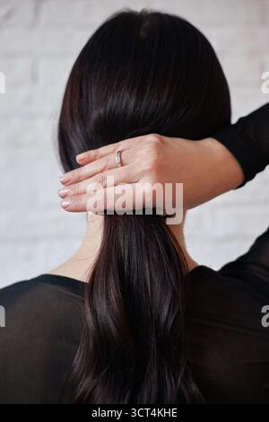 Die Frau wird mit dem Rücken zum Betrachter gesehen, während sie sanft mit einer Hand ihr langes dunkles Haar hält, während sie ein schwarzes Oberteil trägt. Hintergrund mit heller Farbe Stockfoto