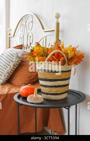 Gewebter Korb gefüllt mit gelben Herbstblättern und kleinem orangefarbenen Kürbis auf schwarzem Tisch neben Bett mit geschmiedetem Kopfteil und Kissen. Herbstdekoration Stockfoto