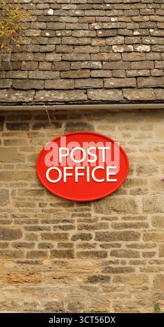 Rotes ovales Postschild mit weißem Text an einer alten rustikalen Steinmauer, das traditionelle Gebäudearchitektur und Postdienstkonzept zeigt. Cotsw Stockfoto