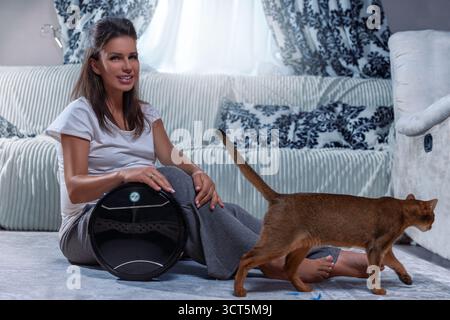 Eine schwangere Frau sitzt bequem auf dem Boden und lächelt, während ein intelligenter Staubsauger den Raum reinigt. Eine Katze spaziert, indem sie der gemütlichen Atmosphäre einen verspielten Touch verleiht. Stockfoto
