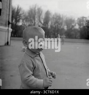 Ein offenes Porträt eines Kleinkindes in einem öffentlichen Park oder Innenhof in Sloviansk der 1960er Jahre, ukrainische SSR. Dieses Schwarzweiß-Foto fängt den unschuldigen und kontemplativen Ausdruck des Kindes ein. Er trägt eine schlichte Strickjacke, typisch für diese Zeit. Im Hintergrund sind Kreideumrisse für ein Hopscotch-Spiel auf dem Asphalt sichtbar, die die Szene in einen Kontext von alltäglichen Straßenspielen und Outdoor-Spielen stellen. Diese charmante und authentische Momentaufnahme eines flüchtigen Moments weckt ein starkes Gefühl der Nostalgie für eine friedliche Kindheit aus der Sowjetzeit Stockfoto