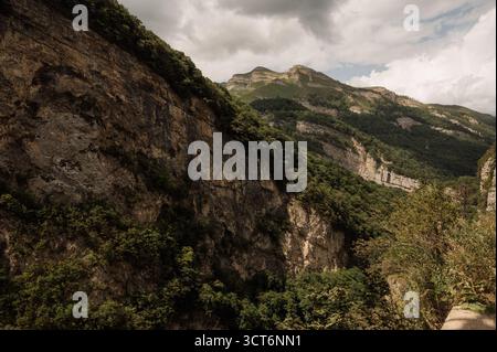 Eine Nahaufnahme einer Felswand mit Vegetation und Berggipfeln tief in einem Canyon. Der steile felsige Hang der Kaukasusschlucht, bedeckt mit dichtem Grün, Stockfoto