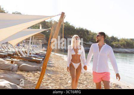Ein junges Paar schlendert Hand in Hand am Sandstrand entlang und taucht in die Sommersonne und lebhafte Küstenatmosphäre ein. Stockfoto