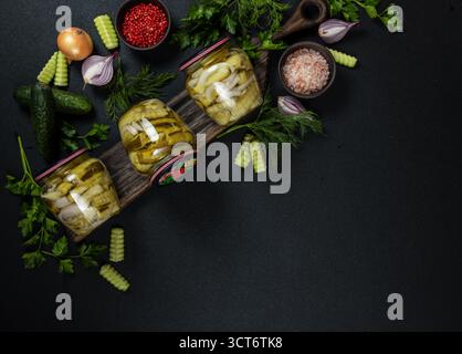 Eingelegte Gurken in Glasgläsern mit frischen Gurken, roten Zwiebeln, Dill und Petersilie auf einem dunklen rustikalen Brett, mit Gewürzen für hausgemachte Konserven angeordnet Stockfoto