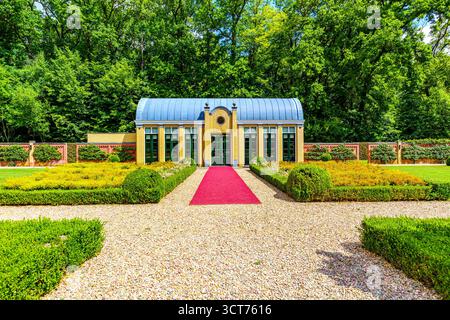 Öffentlicher Park der Burg TerWormista mit Gewächshaus in Frontalansicht, Pfad mit rotem Teppich zum Eingang, Laubbäume im Hintergrund, gelbe Ziegelwände A Stockfoto