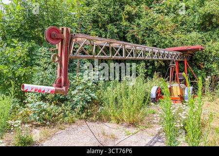 Alter, veralteter Traktor mit einem landwirtschaftlichen Kran, verlassen zwischen üppiger wilder Vegetation des Bauernhofs auf landwirtschaftlichem Boden, rostiger roter Metallarm, Laubbäume Stockfoto