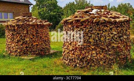 Bauernhof mit kreisförmigen Brennholzpfählen auf grünem Gras, Ziegelmauer und Brennholzregal, kleine Laubbäume im Hintergrund, sonniger Tag in Limburg, Belgiu Stockfoto