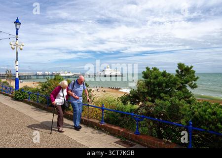 Ein älteres Ehepaar, das an einem sonnigen Sommertag in East Sussex England UK mit Wanderstöcken entlang der Küste von Eastbourne spaziert, mit dem Pier im Hintergrund Stockfoto