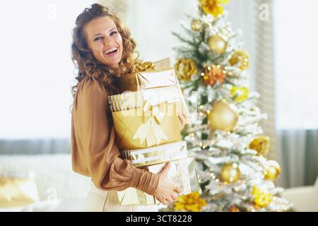 Eine lächelnde Frau mit lockigen Haaren hält einen Stapel wunderschön gewickelter Weihnachtsgeschenke. Hinter ihr leuchtet ein mit goldenen Ornamenten geschmückter Baum sanft unter Stockfoto